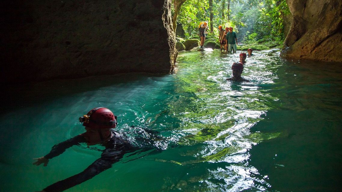 A WeRoad group trip wearing helmets swims through a turquoise river at the entrance of a jungle cave.