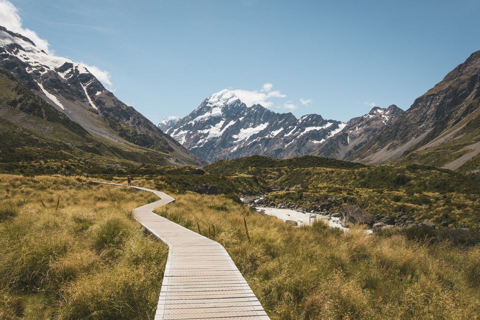 A wooden boardwalk winds through a grassy valley towards towering, snow-capped mountains under a clear blue sky.