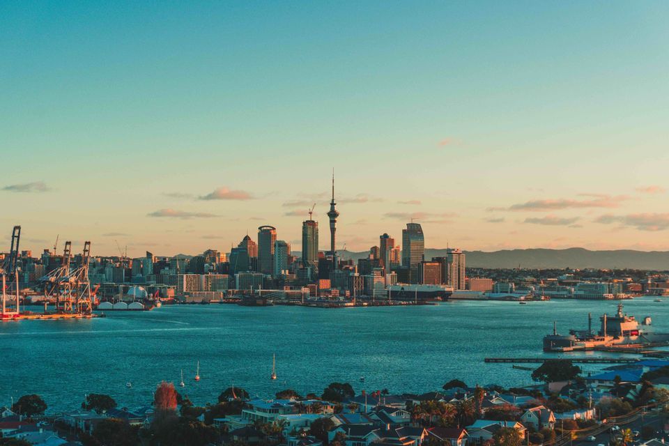 A city skyline with a tall central tower sits across a body of water at sunset, with a port and sailboats in view.