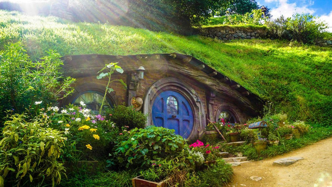 A house with a round blue door built into a green hillside, surrounded by a colorful garden under bright sunlight.