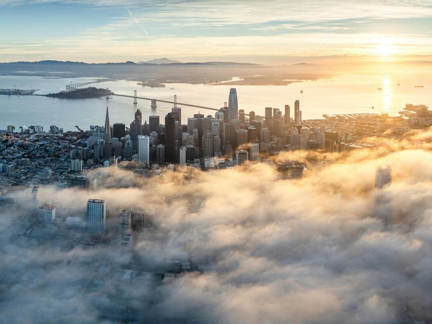 Aerial view of a city skyline with skyscrapers emerging from a dense blanket of fog as the sun rises over the water.