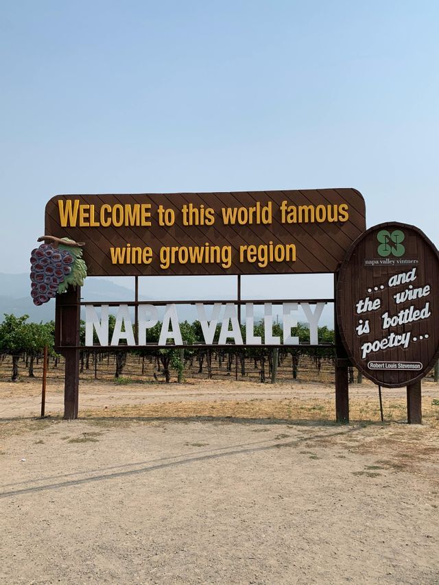 A large wooden welcome sign for Napa Valley, a wine growing region, stands in front of a vineyard with rows of grapevines.