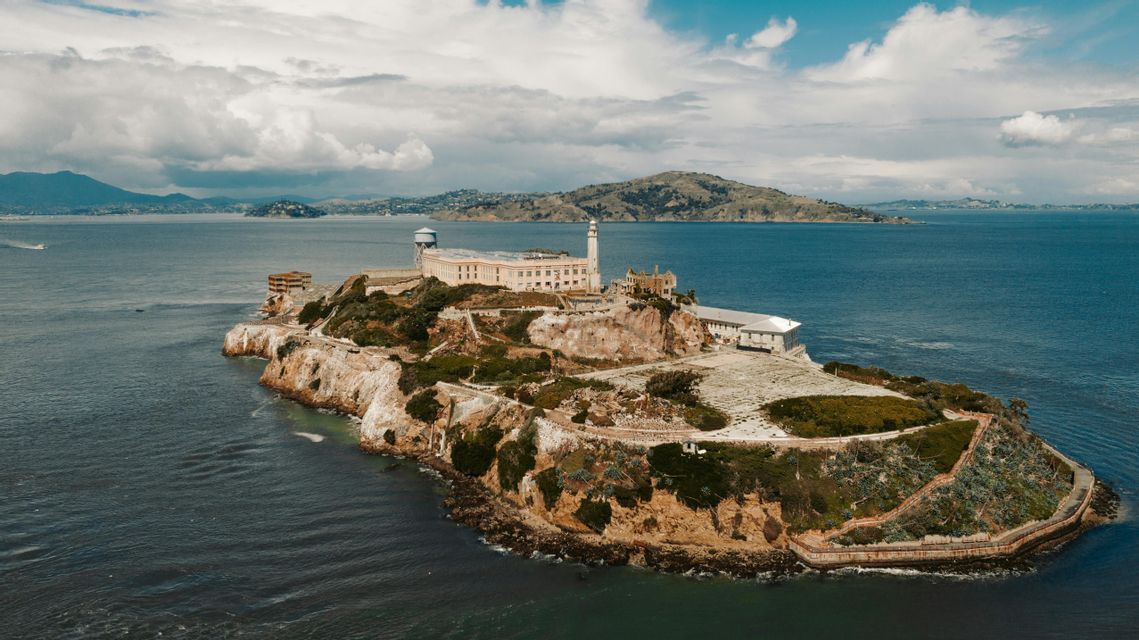 An aerial view of a historic prison complex on a rocky island, surrounded by water with distant hills under a cloudy sky.