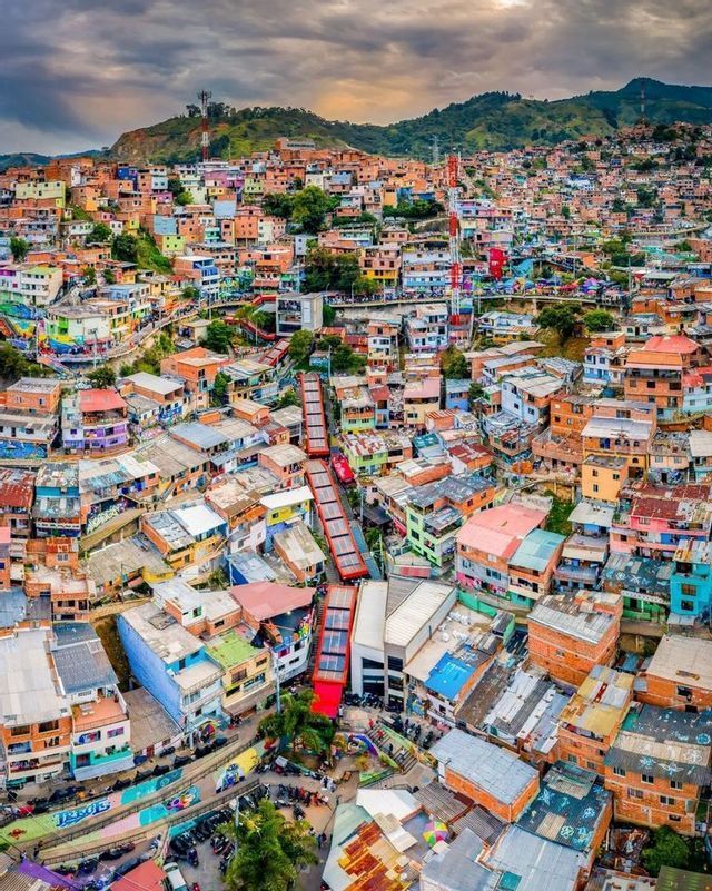 An aerial view of red outdoor escalators ascending a hillside through a dense, colorful neighborhood under a cloudy sky.