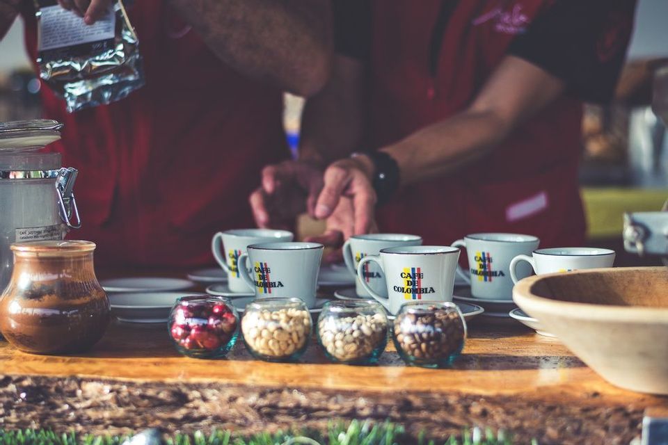 A close-up of a coffee tasting with bowls of coffee beans and mugs that say 'Cafe de Colombia' on a wooden surface.
