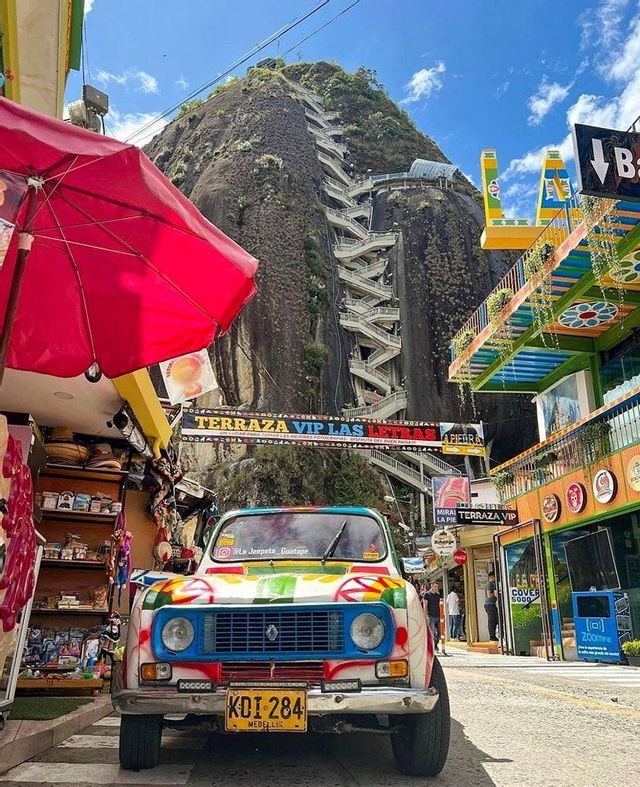 A colorful vintage car parked on a street below a large rock mountain with a prominent zig-zagging staircase leading up its side.