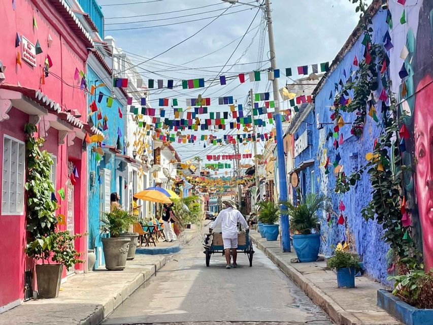 A man pushes a cart down a narrow street lined with colorful buildings and decorated with overhead flags.