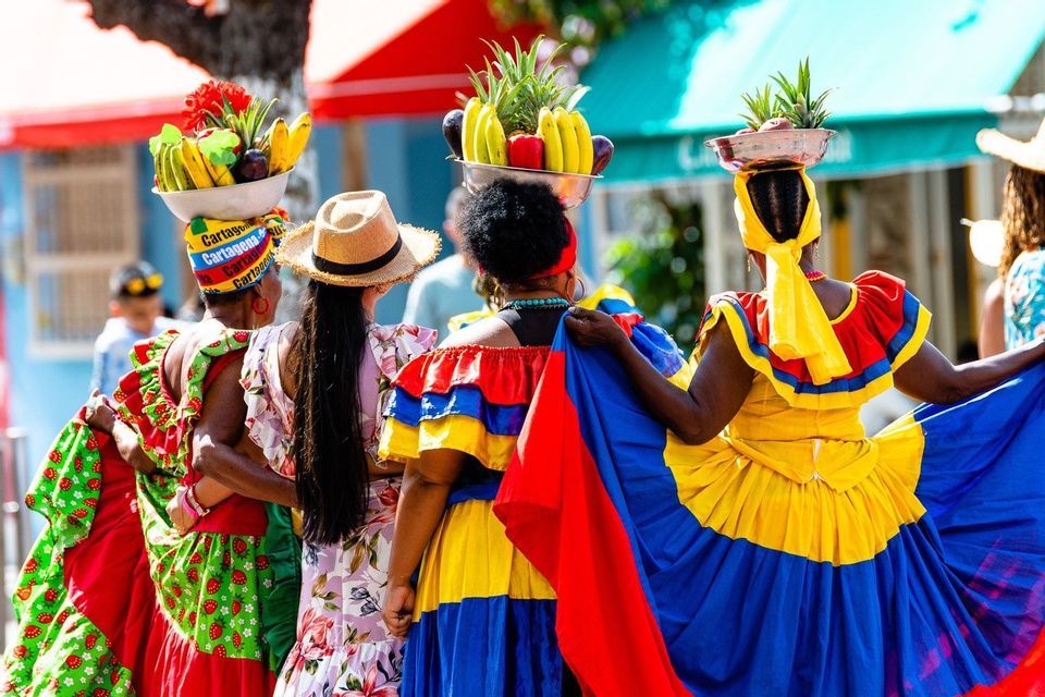 A group of women seen from behind in colorful, traditional dresses, balancing bowls of fresh fruit on their heads.