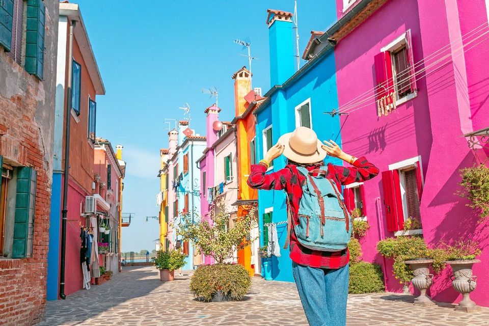 Una persona con uno zaino, vista da dietro, si tiene il cappello mentre è in piedi su una strada fiancheggiata da edifici colorati.