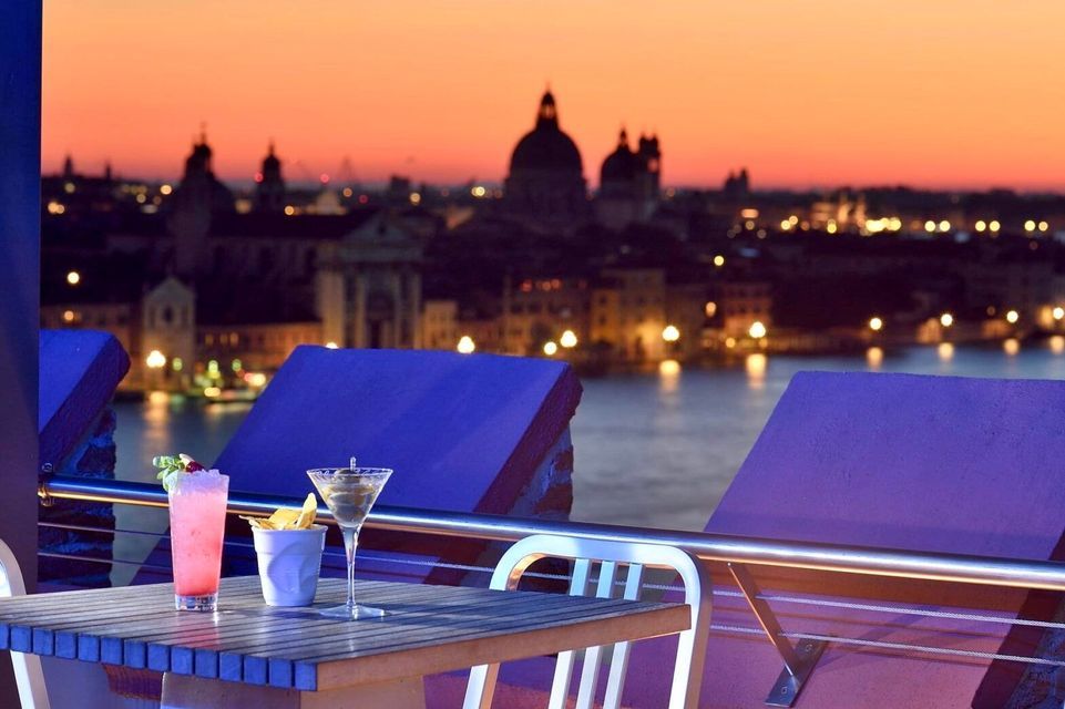 Due cocktail su un tavolo in un bar panoramico con vista sullo skyline illuminato della città e sull'acqua al tramonto.