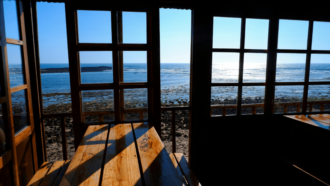 Un tavolo di legno in una stanza buia, posizionato accanto a grandi finestre a riquadri, offre una vista soleggiata su una costa rocciosa e sull'oceano.