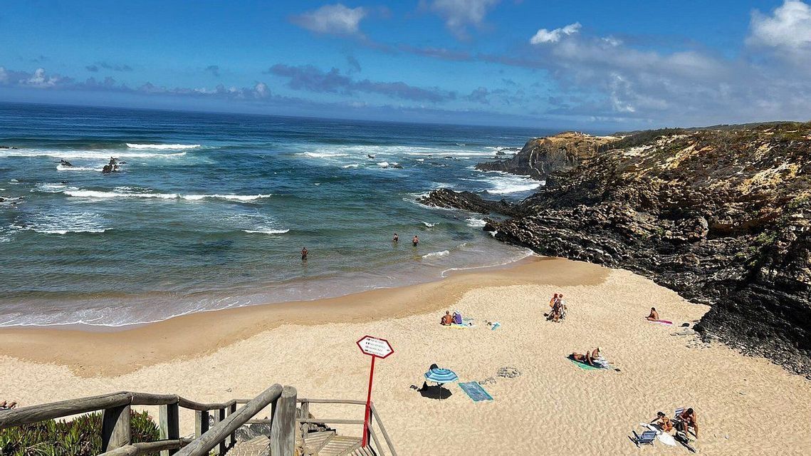 Una vista dall'alto di una spiaggia sabbiosa in una caletta rocciosa, con persone che nuotano nel mare e prendono il sole sulla sabbia.