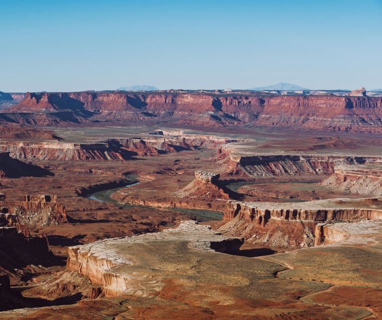 An expansive view of a deep red rock canyon with a river snaking through the canyon floor under a clear sky.