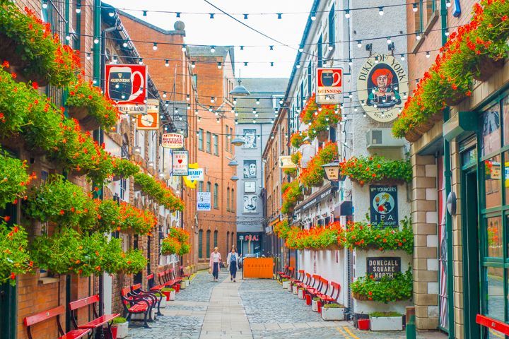 Un vicolo stretto e acciottolato, fiancheggiato da edifici in mattoni, insegne di pub, cesti di fiori appesi e lucine.