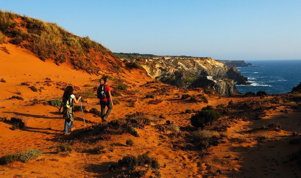 Due escursionisti con zaini percorrono un sentiero costiero sabbioso e arancione, con scogliere a picco sull'oceano.