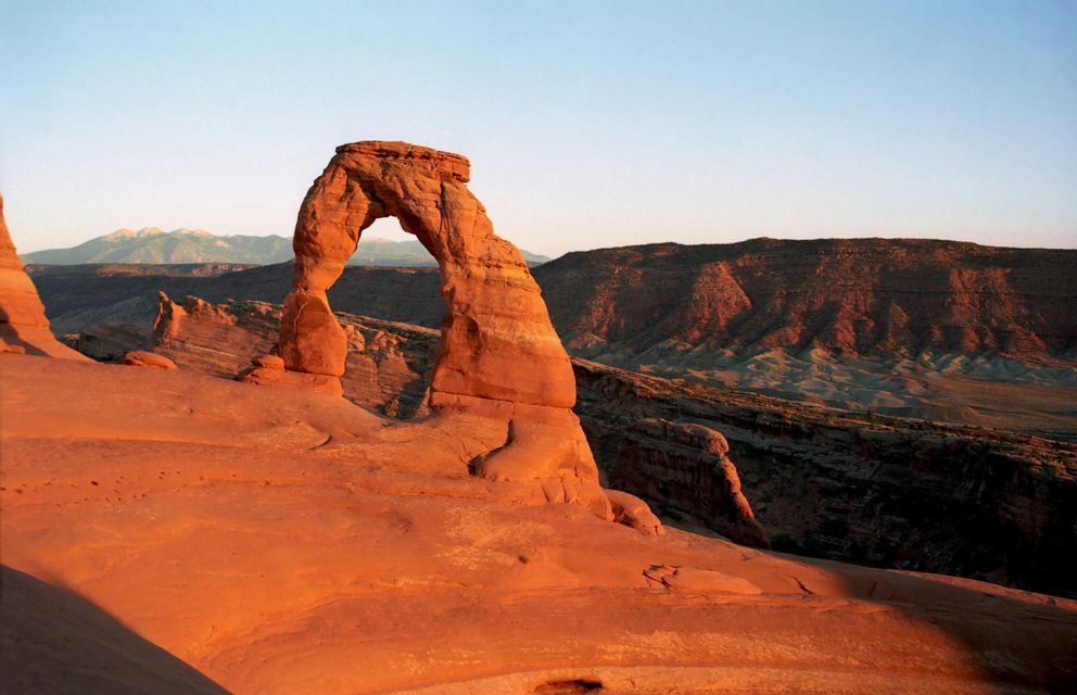 A large, natural sandstone arch glows in the warm light of sunrise over a rocky desert landscape with mountains in the distance.