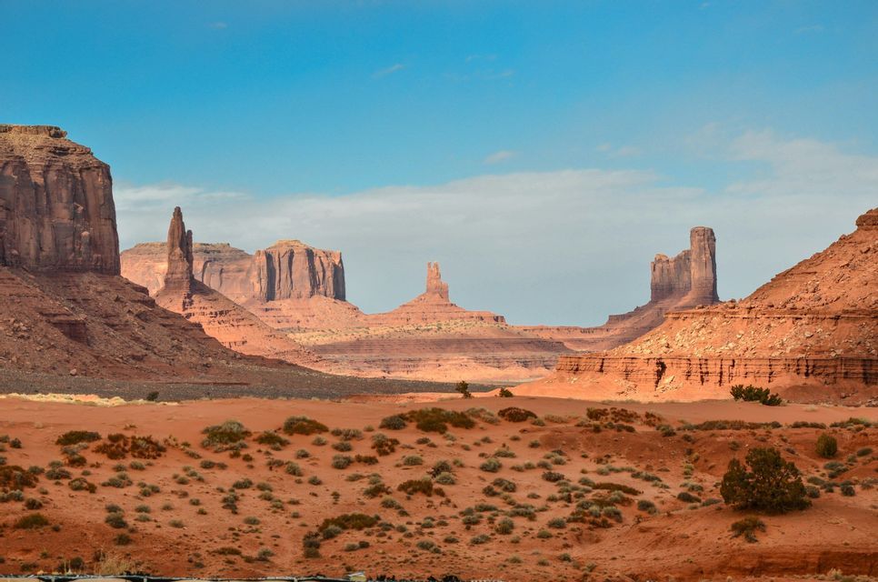 A wide shot of a red sand desert valley with large sandstone buttes and mesas rising in the distance under a blue sky.
