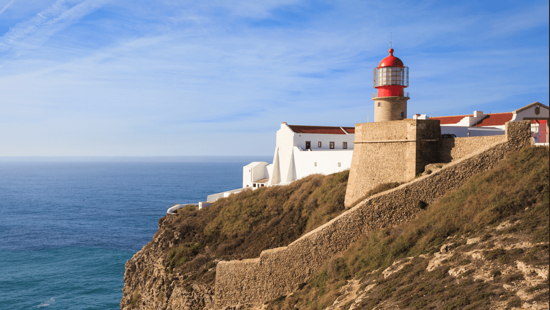 Un faro di pietra con la sommità rossa e edifici bianchi adiacenti si ergono su una scogliera rocciosa a picco sull'oceano sotto un cielo azzurro.