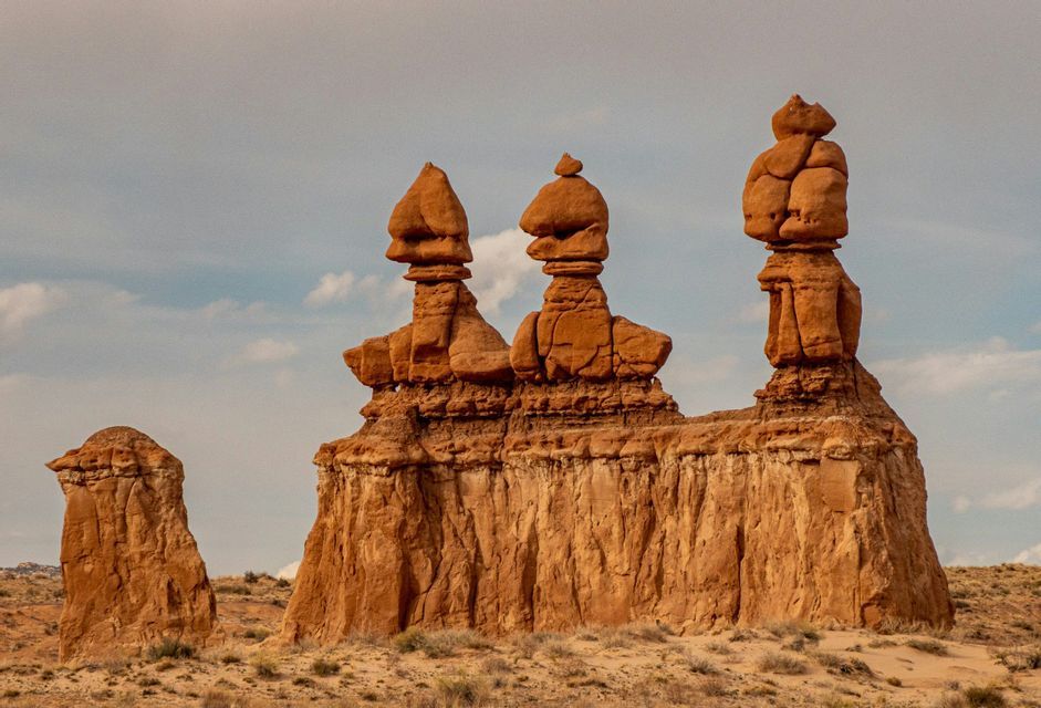Three large, reddish-brown hoodoo rock formations stand in a desert landscape under a partly cloudy sky.