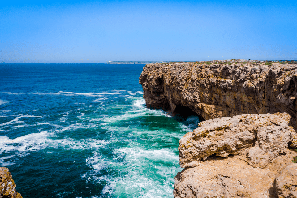 Scogliere aspre color ocra dominano un mare turchese e azzurro impetuoso con onde bianche che si infrangono sotto un cielo sereno.