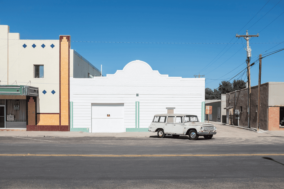 A vintage white station wagon parked on a street in front of minimalist storefronts under a clear blue sky.
