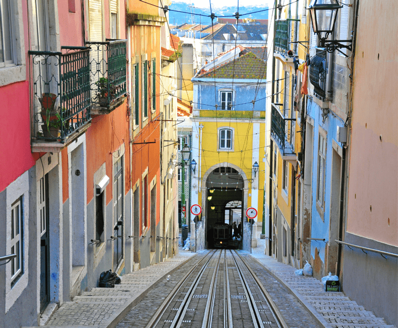 Una vista in discesa su una ripida strada acciottolata con binari del tram, fiancheggiata da edifici colorati e con una funicolare in fondo.