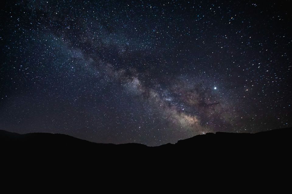 The Milky Way galaxy arcs across a dark, star-filled night sky above the silhouette of mountains.