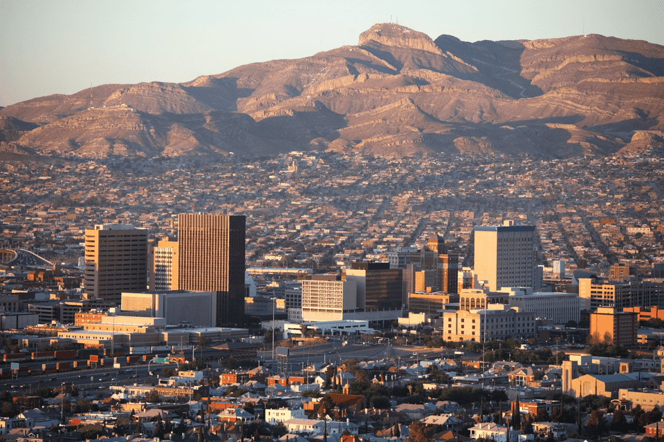 An expansive city skyline with downtown buildings is nestled at the base of a large, arid mountain range at golden hour.
