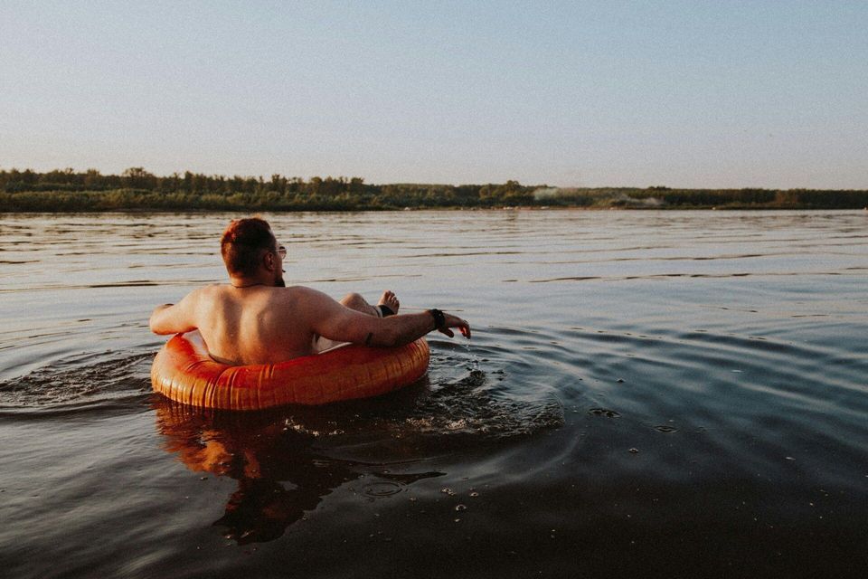 A shirtless man seen from behind relaxes in an orange inflatable ring on calm water at sunset.