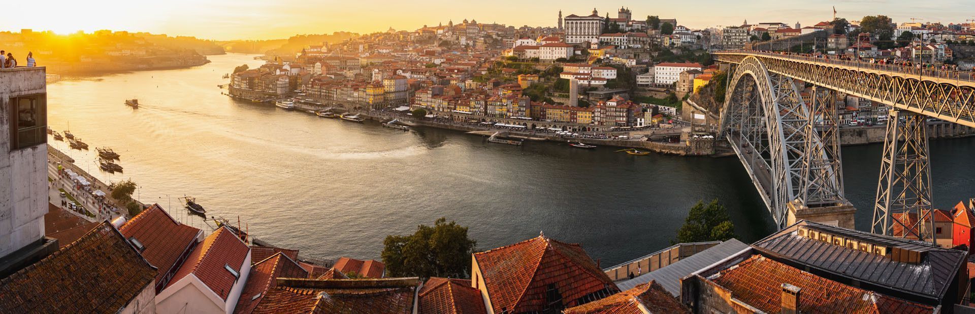 Una vista panoramica di una città sul fiume con un grande ponte di metallo e edifici colorati illuminati dalla luce dorata del tramonto.