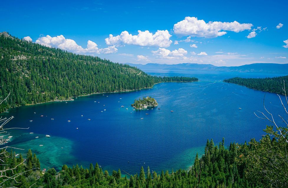 A high-angle view of a deep blue lake with boats surrounding a small, tree-covered island, all enclosed by forested hills under a blue sky.