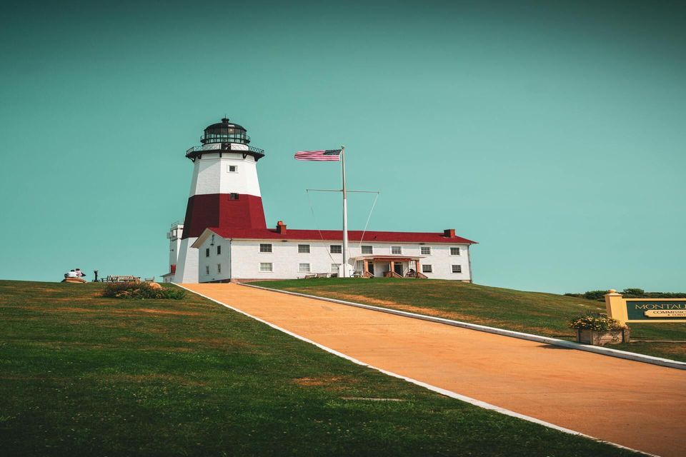 A red and white lighthouse with an adjacent building sits atop a grassy hill under a clear teal sky, with a path leading up to it.