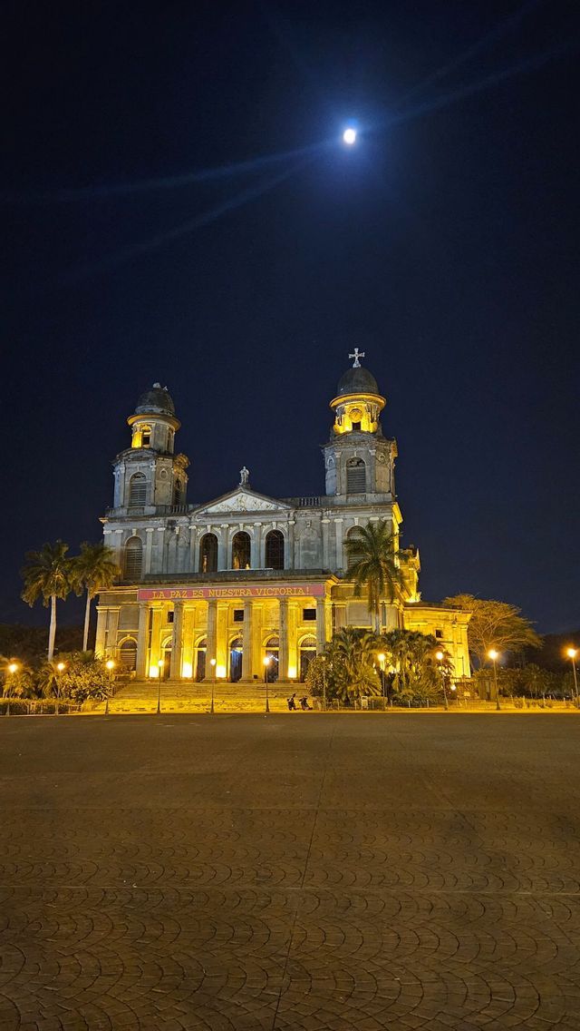 Eine große, historische Kathedrale ist nachts beleuchtet, davor ein weiter, leerer Kopfsteinpflasterplatz, vor einem dunklen Himmel mit hellem Mond.