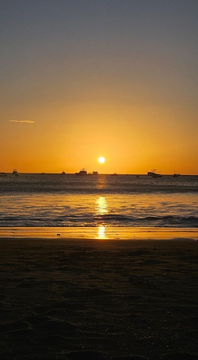 Die Sonne versinkt über dem Meer, Boote zeichnen sich als Silhouetten am Horizont ab, während sich ihr Schein auf dem nassen Sand spiegelt.