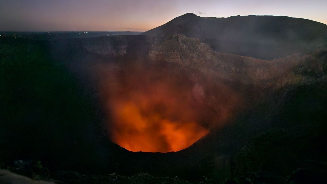 Weitblick auf einen Vulkankrater bei Sonnenuntergang, mit glühender orangefarbener Lava und aufsteigendem Rauch.