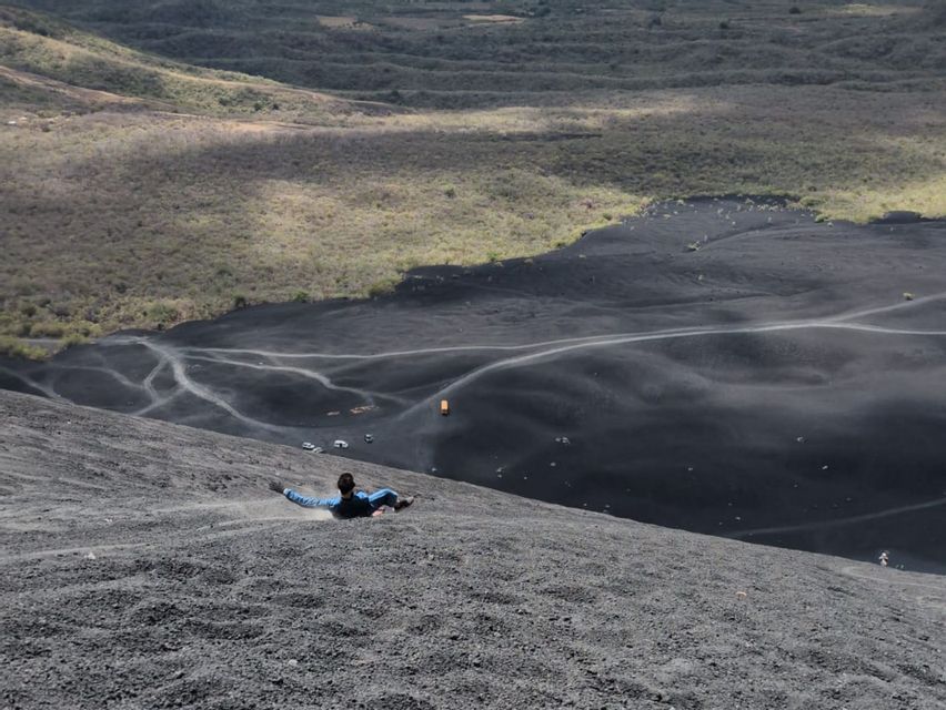 Eine Person rutscht einen steilen Hang aus schwarzem Vulkangestein hinab, mit Blick auf ein großes Tal darunter.