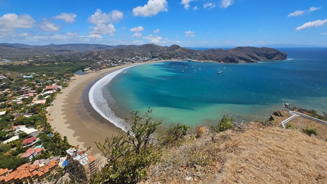 Vogelperspektive auf eine Küstenstadt an einem geschwungenen Sandstrand, angrenzend an eine türkisfarbene Bucht mit Booten, unter einem teils bewölkten Himmel.