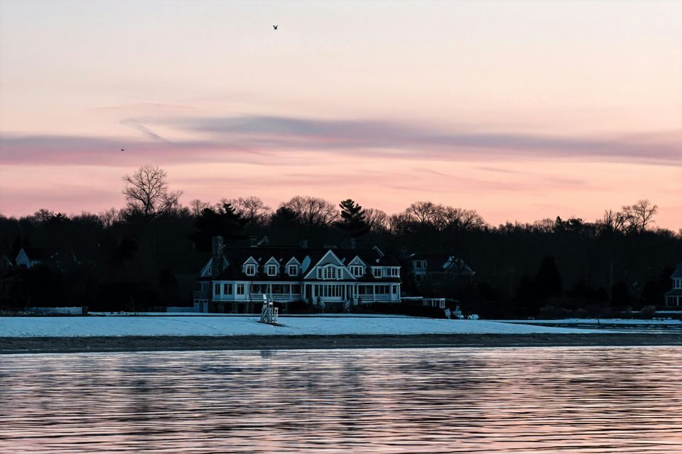 A large white house sits on a snowy shoreline across from a body of water, under a pink and purple sunset sky.