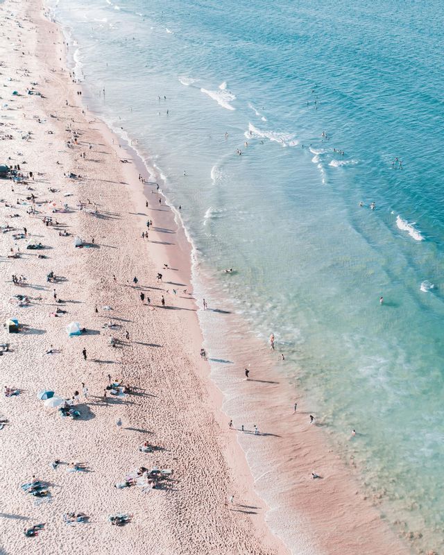 An aerial view of many people relaxing on a sandy beach and swimming in the turquoise ocean.