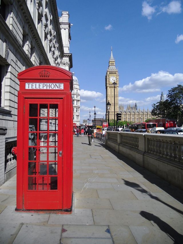 A red telephone booth on a sidewalk with the Big Ben clock tower in the background on a sunny day.