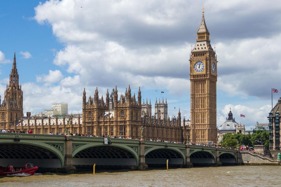 A large clock tower and a historic building stand behind a crowded stone bridge over a river under a partly cloudy sky.