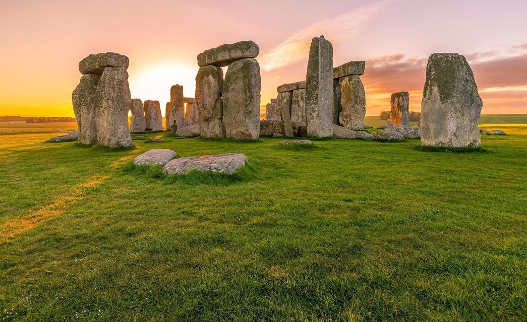 The sun sets behind a circle of ancient standing stones on a lush green field.