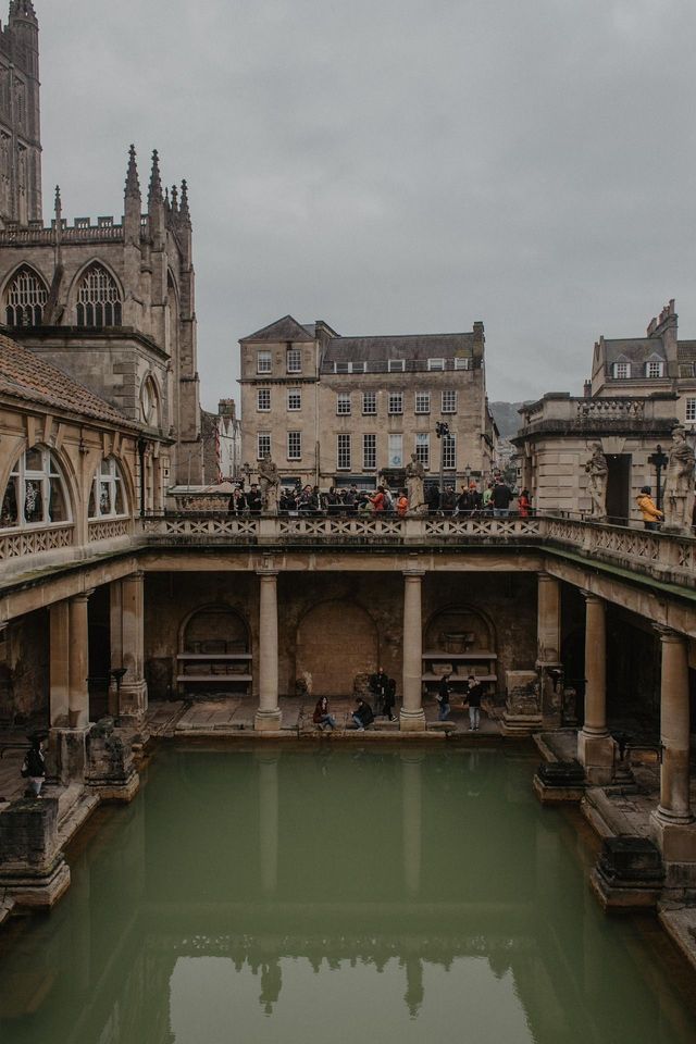 People stand on a terrace overlooking a large thermal pool surrounded by historic stone architecture under a cloudy sky.