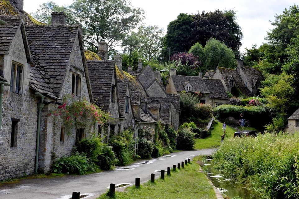 A row of historic stone cottages with slate roofs lines a narrow road next to a small stream in a lush, green village.