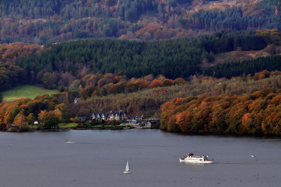 A large white ferry and a sailboat on a lake with a shoreline of buildings and autumn-colored forests.