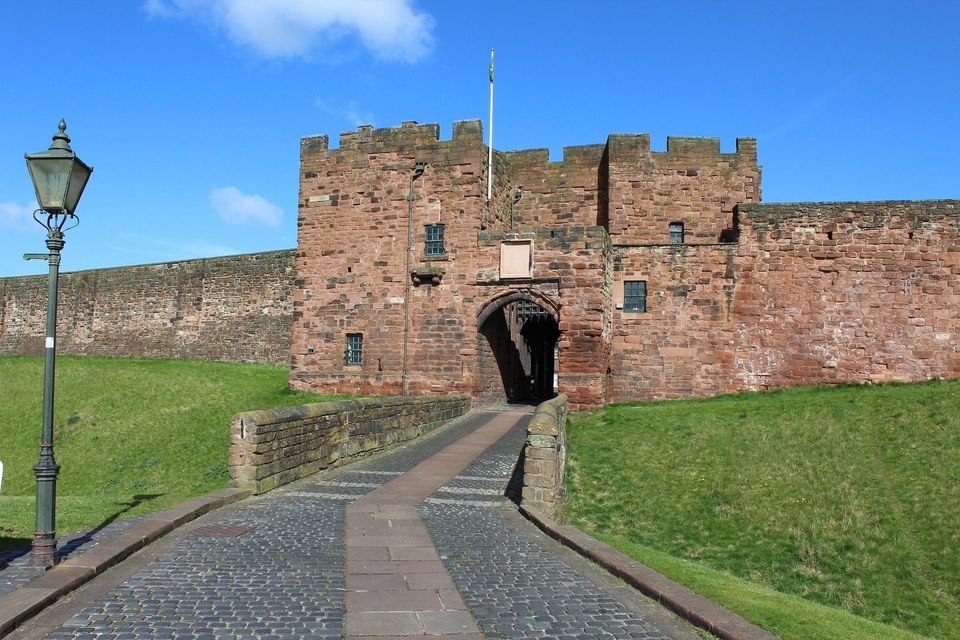 A cobblestone path leads over a bridge to the gatehouse of a large red stone castle under a clear blue sky.