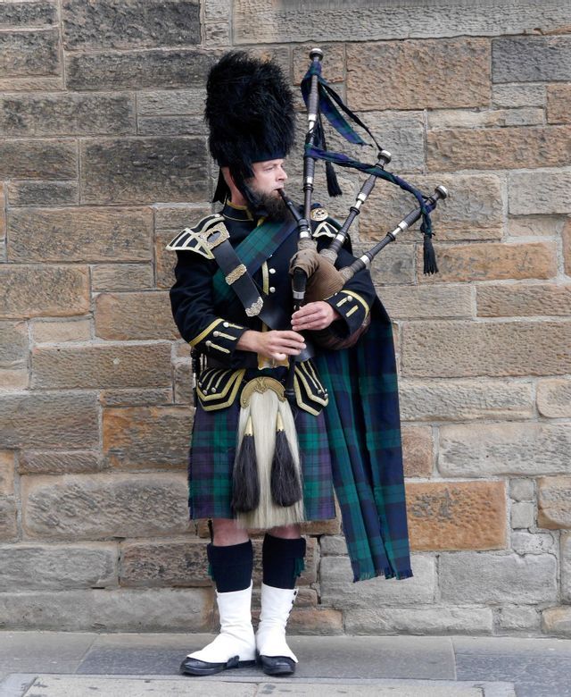 A man in traditional Scottish dress, including a tartan kilt and tall bearskin hat, plays the bagpipes in front of a stone wall.