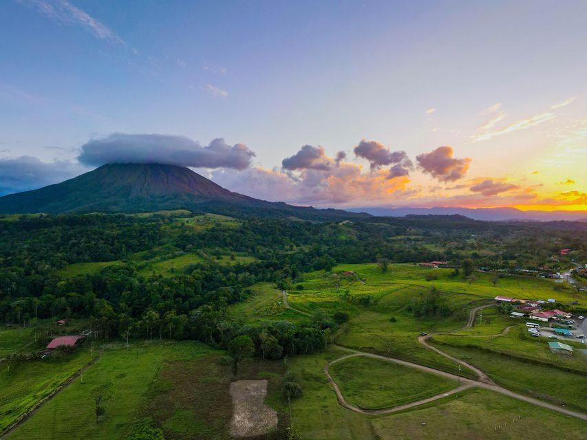 Una vista aerea di un vulcano con la sua cima tra le nuvole, che domina una valle verde lussureggiante e una piccola città al tramonto.