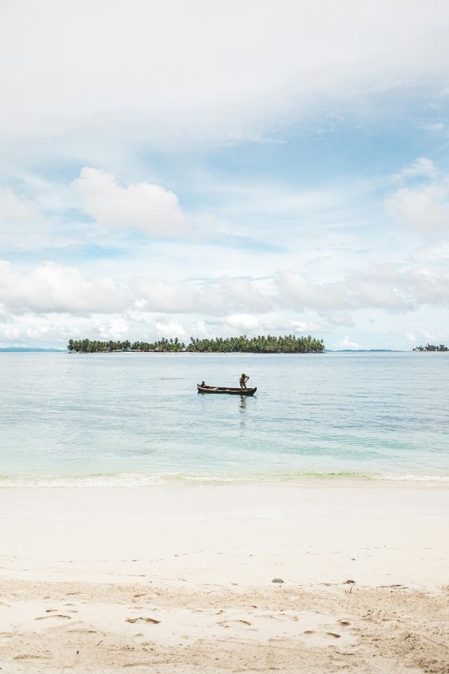 Una persona pagaia una piccola canoa su acque calme al largo di una spiaggia di sabbia bianca, con un'isola ricoperta di palme all'orizzonte.