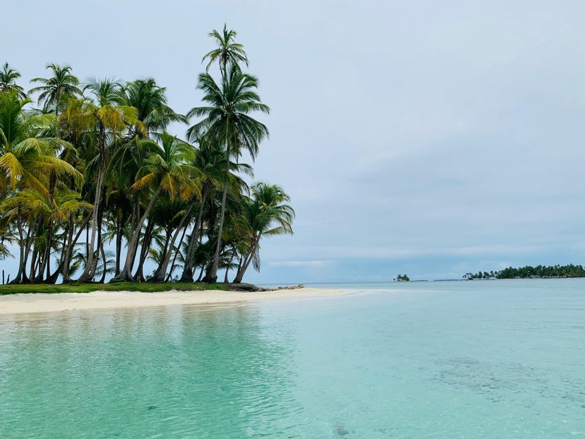 Una piccola isola tropicale con una spiaggia di sabbia bianca e molte palme, circondata da acque turchesi e cristalline sotto un cielo nuvoloso.